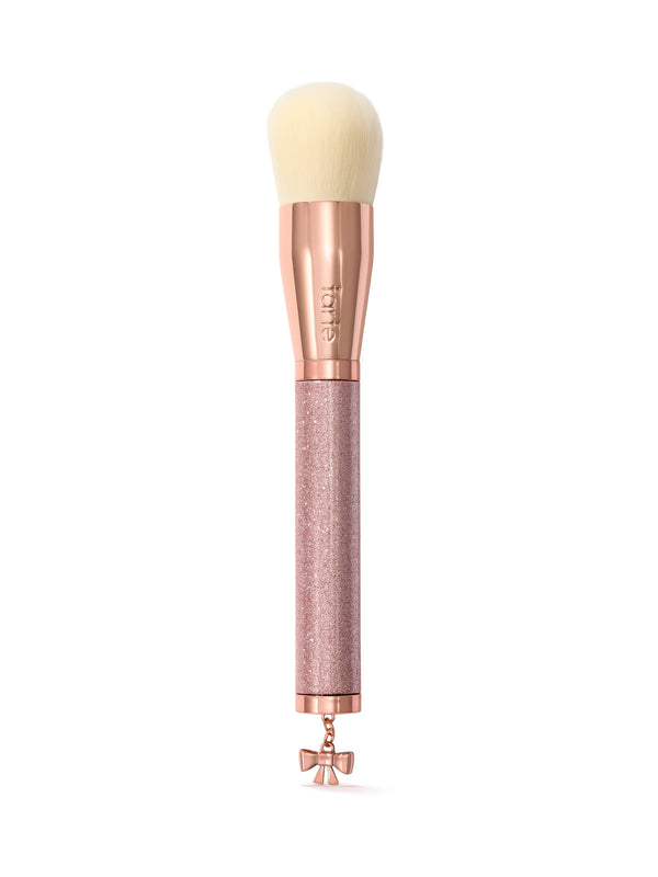 A makeup brush with cream-colored bristles, a rose gold ferrule, and a glittery rose gold handle featuring a small bow charm at the tip. The brand name 'tarte' is visible on the ferrule.