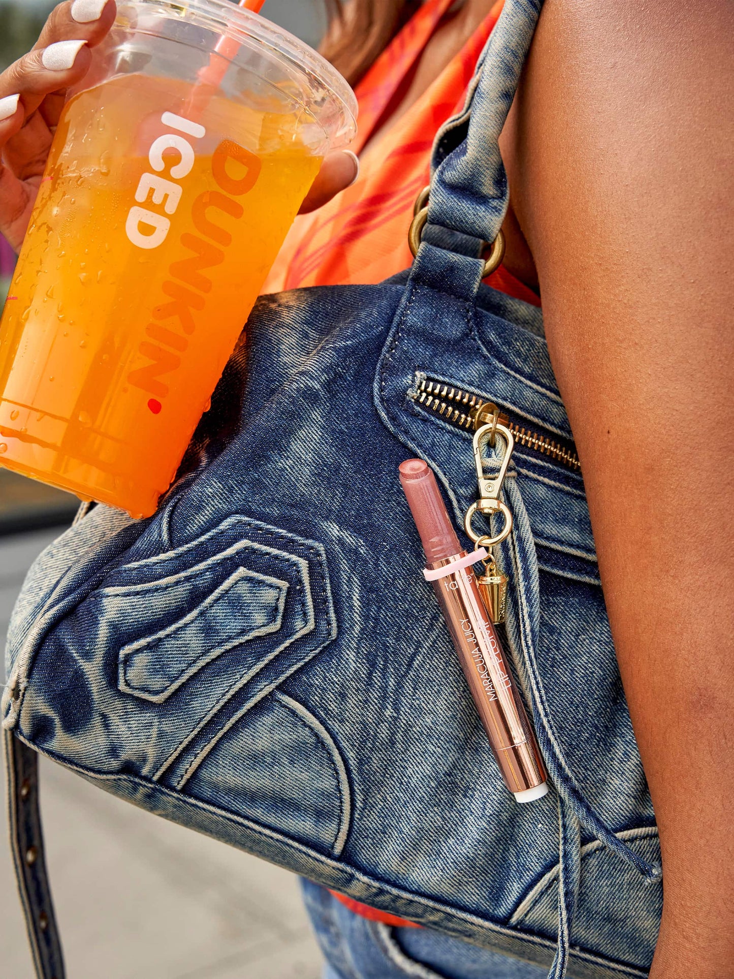 Person holding a Dunkin' iced coffee cup with a denim bag and pink lip gloss.
