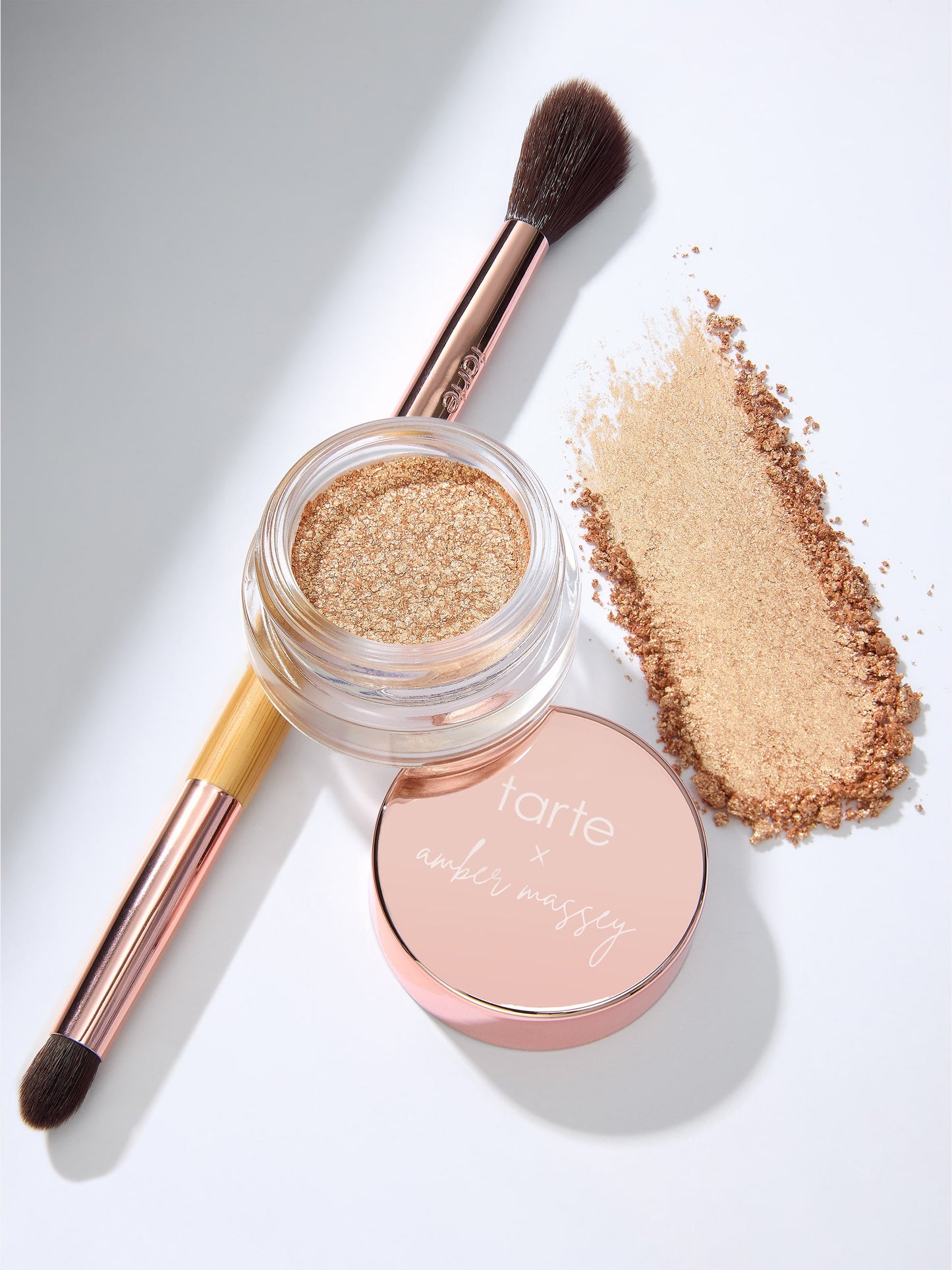 A makeup brush rests beside an open glittery gold eyeshadow pot, with a matching lid reading tarte x amber massey, on a white surface.