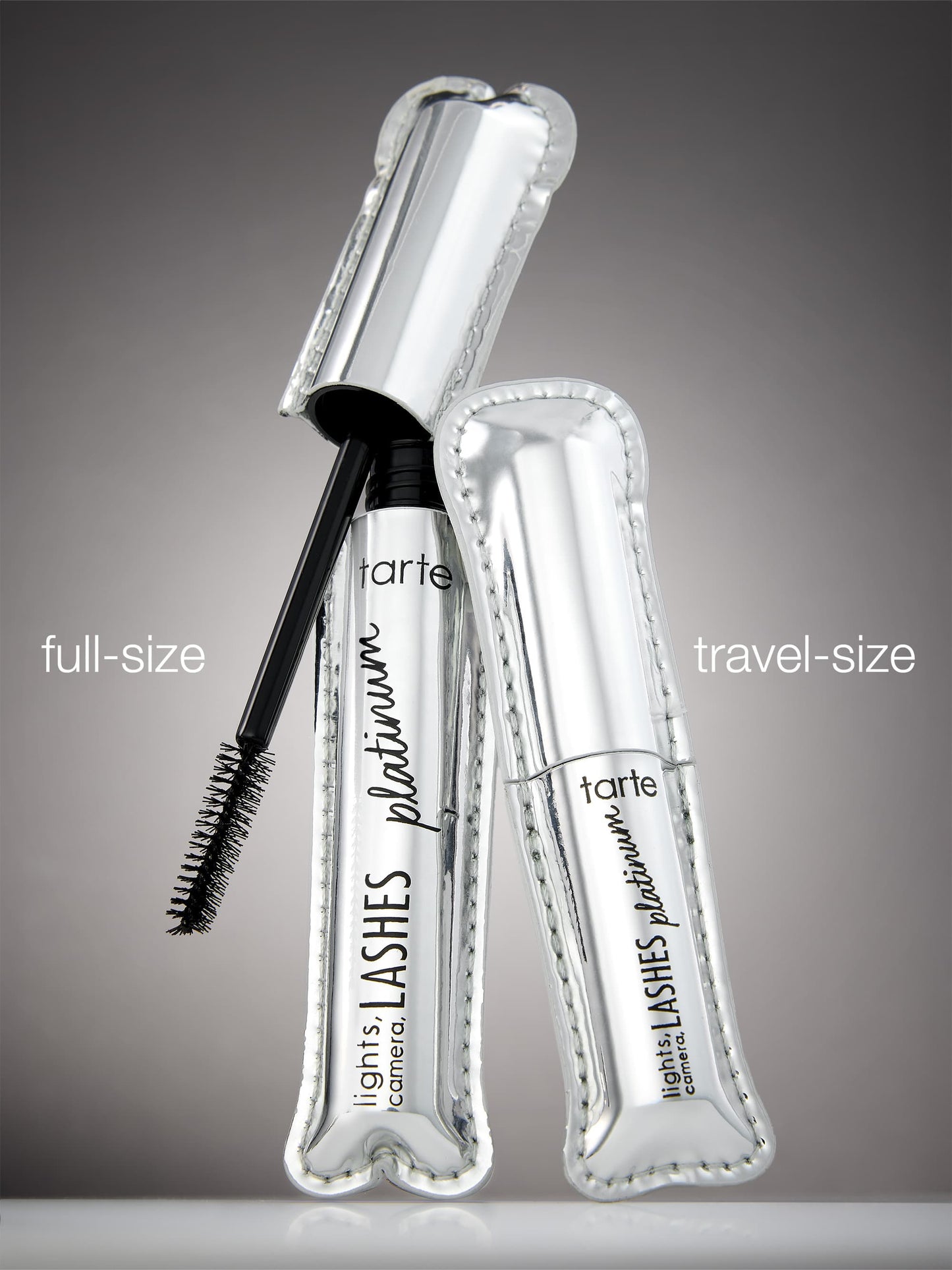 Two shiny silver mascara tubes labeled Tarte lights, camera, lashes platinum stand upright. One tube is open, showing the brush. The text full-size and travel-size appears above them against a gray background.