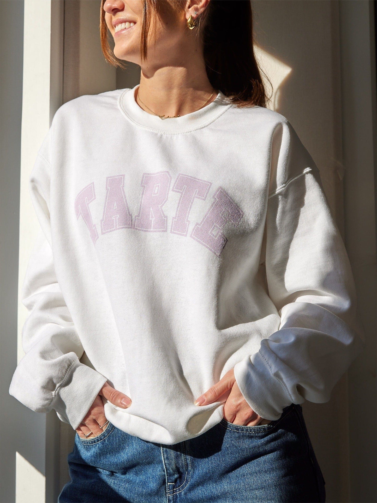 A person wearing a white crewneck sweatshirt with the brand name 'Tarte' written across the front in pink letters.