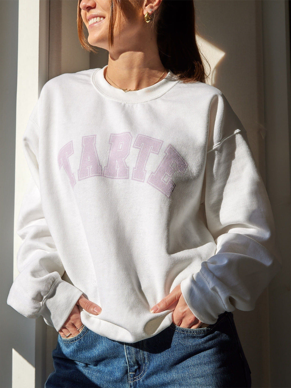 A person wearing a white crewneck sweatshirt with the brand name 'Tarte' written across the front in pink letters.
