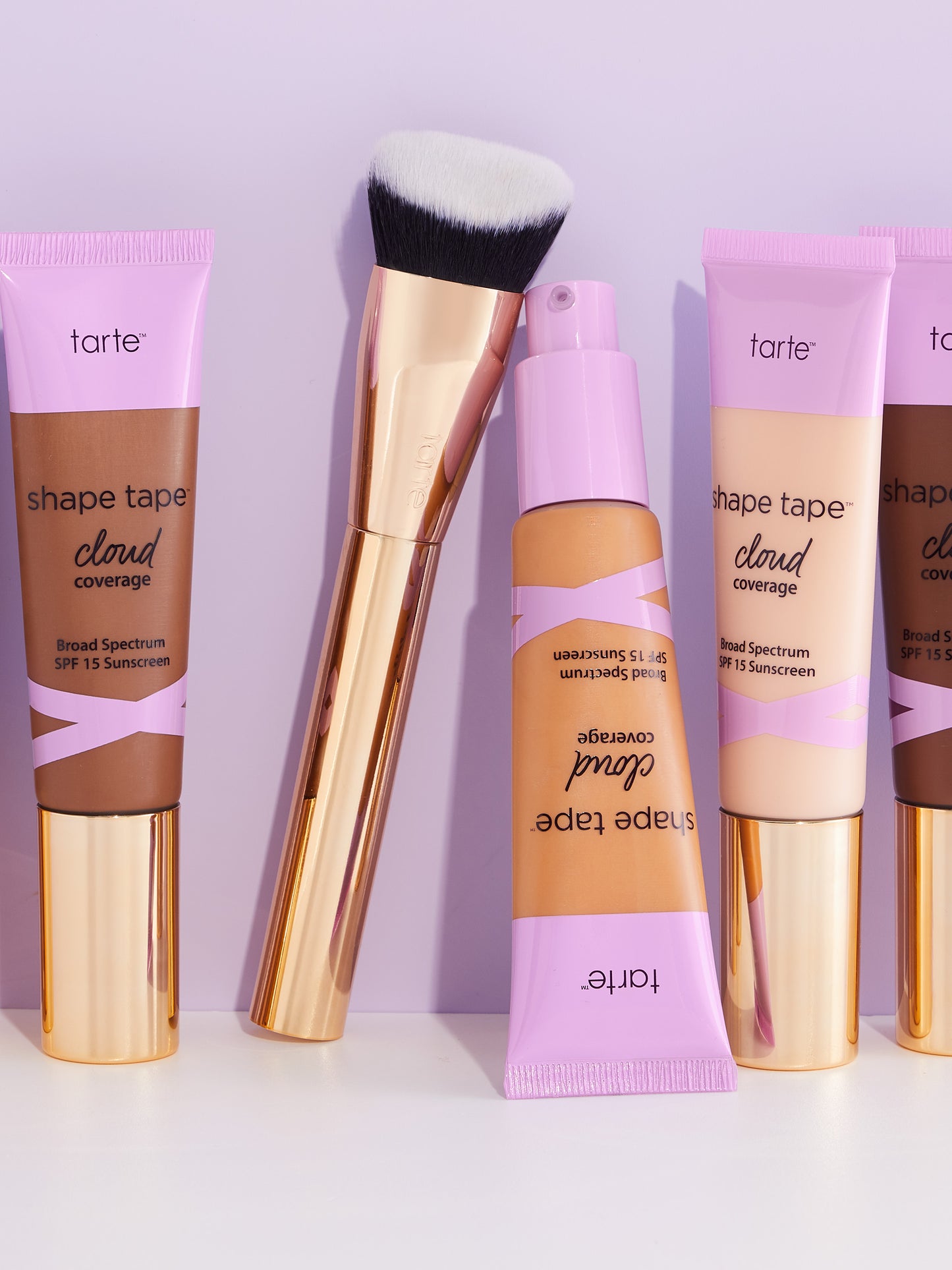 Tarte Cosmetics products stand upright on a white surface. A makeup brush leans against a bottle. Tubes labeled shape tape cloud coverage Broad Spectrum SPF 15 Sunscreen display various shades.