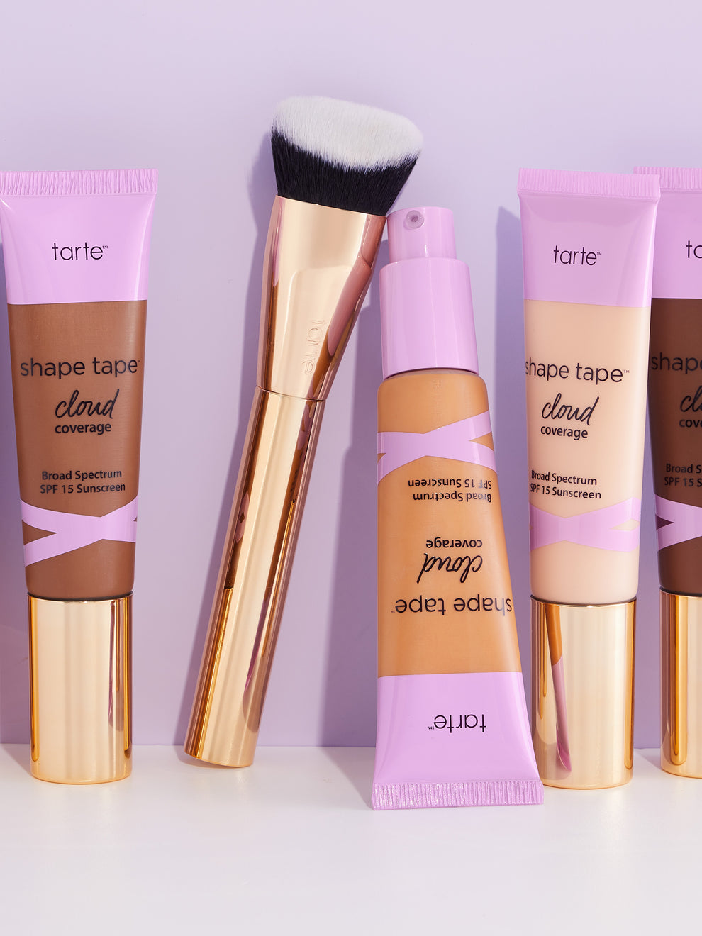 Tarte Cosmetics products stand upright on a white surface. A makeup brush leans against a bottle. Tubes labeled shape tape cloud coverage Broad Spectrum SPF 15 Sunscreen display various shades.