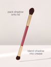 A dual-ended makeup brush with brown bristles, labeled for packing shadow onto the lid and blending into the crease, is displayed against a neutral background. Text: pack shadow onto lid, blend shadow into crease.