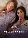 Two women in matching pajamas lying in bed, applying tarte maracuja juicy lip vinyl and looking at a phone. tarte cosmetics x roller rabbit collaboration