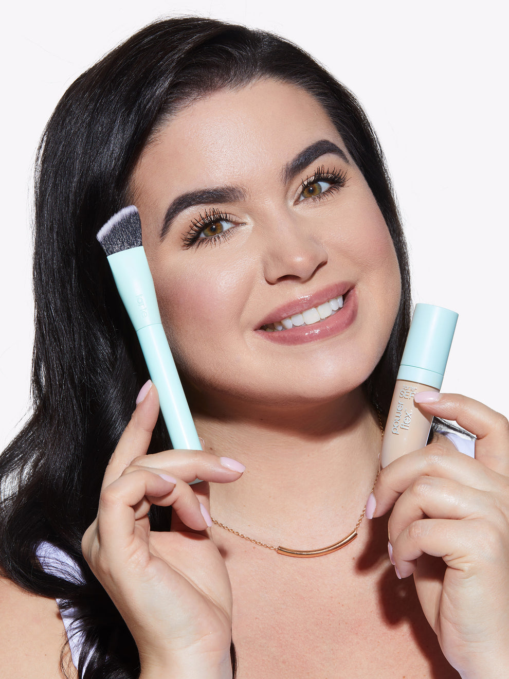 A person smiling holds a turquoise-handled makeup brush and a Tarte Cosmetics Power Flex foundation bottle against a plain white background.