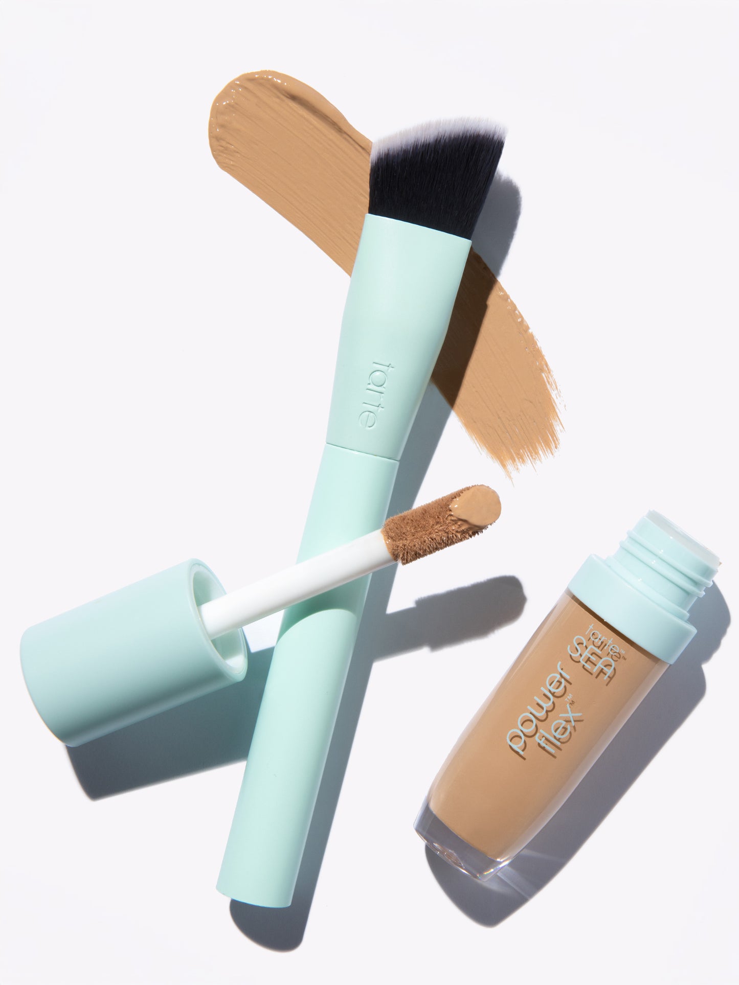 Makeup brush stands beside opened concealer with brush applicator, both featuring mint green handles. Beige makeup swatches beneath suggest application. Text: tarte on brush; power flex water SEA on concealer bottle.
