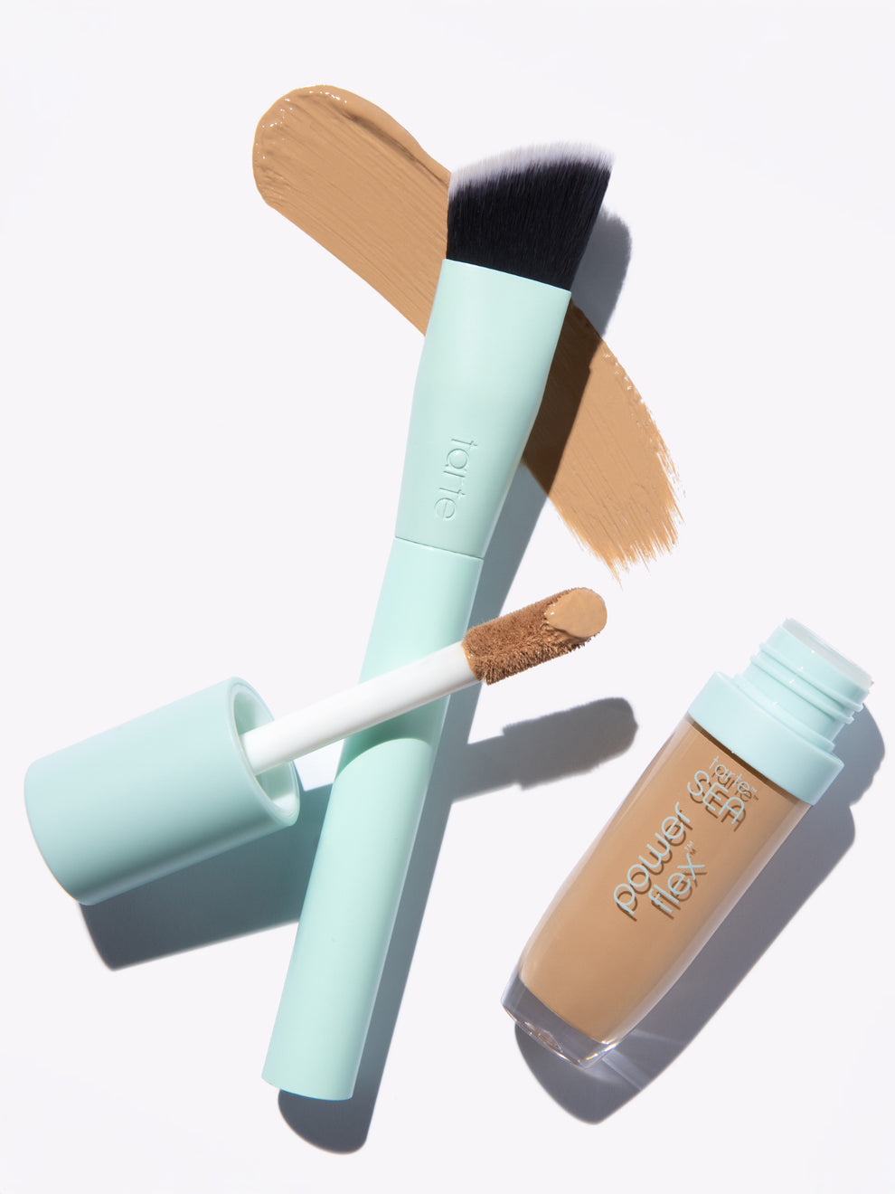 Makeup brush stands beside opened concealer with brush applicator, both featuring mint green handles. Beige makeup swatches beneath suggest application. Text: tarte on brush; power flex water SEA on concealer bottle.