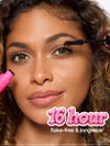 Close-up of a woman applying mascara to her upper lashes, with a bright pink mascara tube visible at the lower left. Text on the image reads '16-hr flake-free & longwear.
