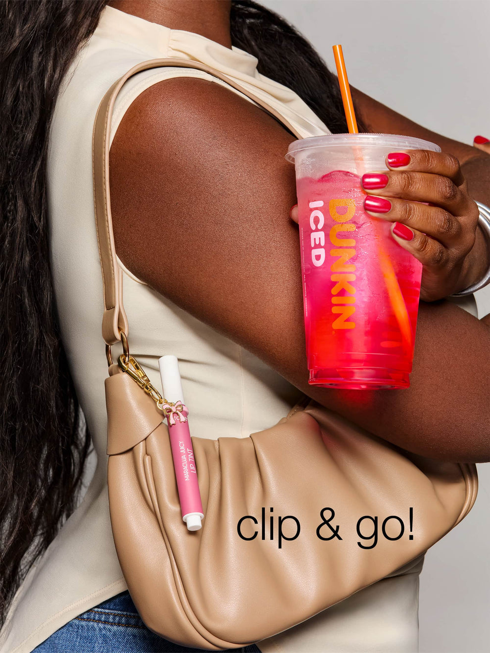 A hand holding an iced drink with pink liquid, while the other arm holds a beige purse featuring a clipped Tarte Cosmetics item. Text reads clip & go!