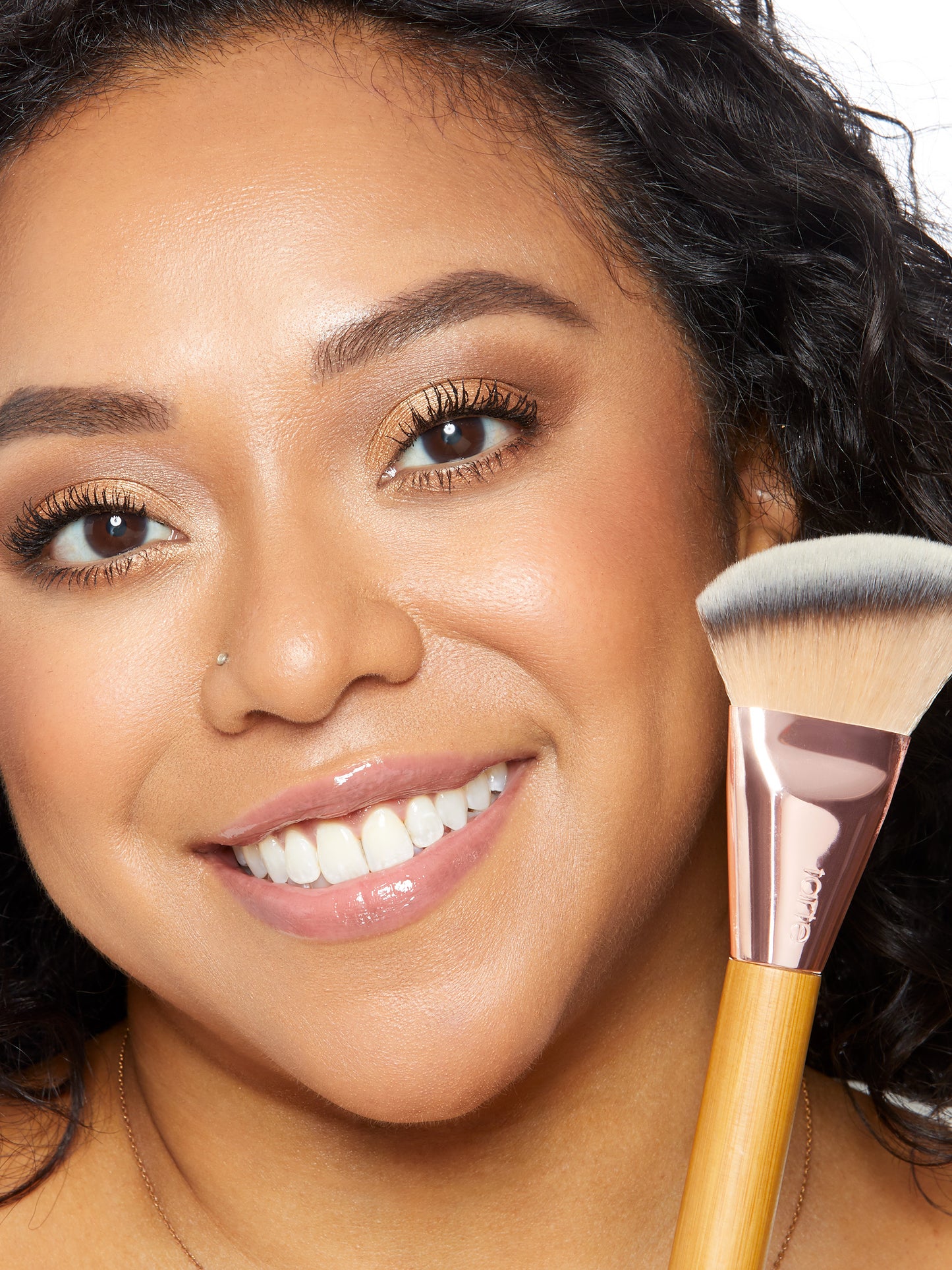 A smiling person holds up a makeup brush with tarte written on the handle, showcasing natural makeup in a bright, well-lit setting.