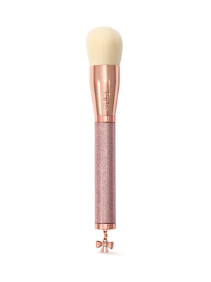 A makeup brush with cream-colored bristles, a rose gold ferrule, and a glittery rose gold handle featuring a small bow charm at the tip. The brand name 'tarte' is visible on the ferrule.