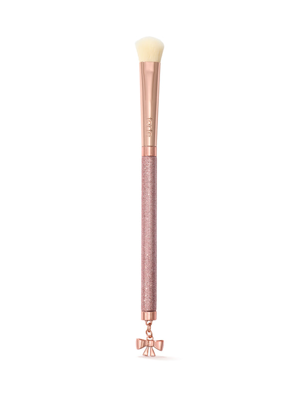 A makeup brush with a rose gold handle, a textured pink glitter grip, and a dangling bow charm at the bottom, shown on a clean white background.