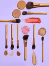 Makeup brushes with wooden handles are laid out with various cosmetic powders and creams, forming a neat arrangement on a lavender background. Text on the brushes: tarte.