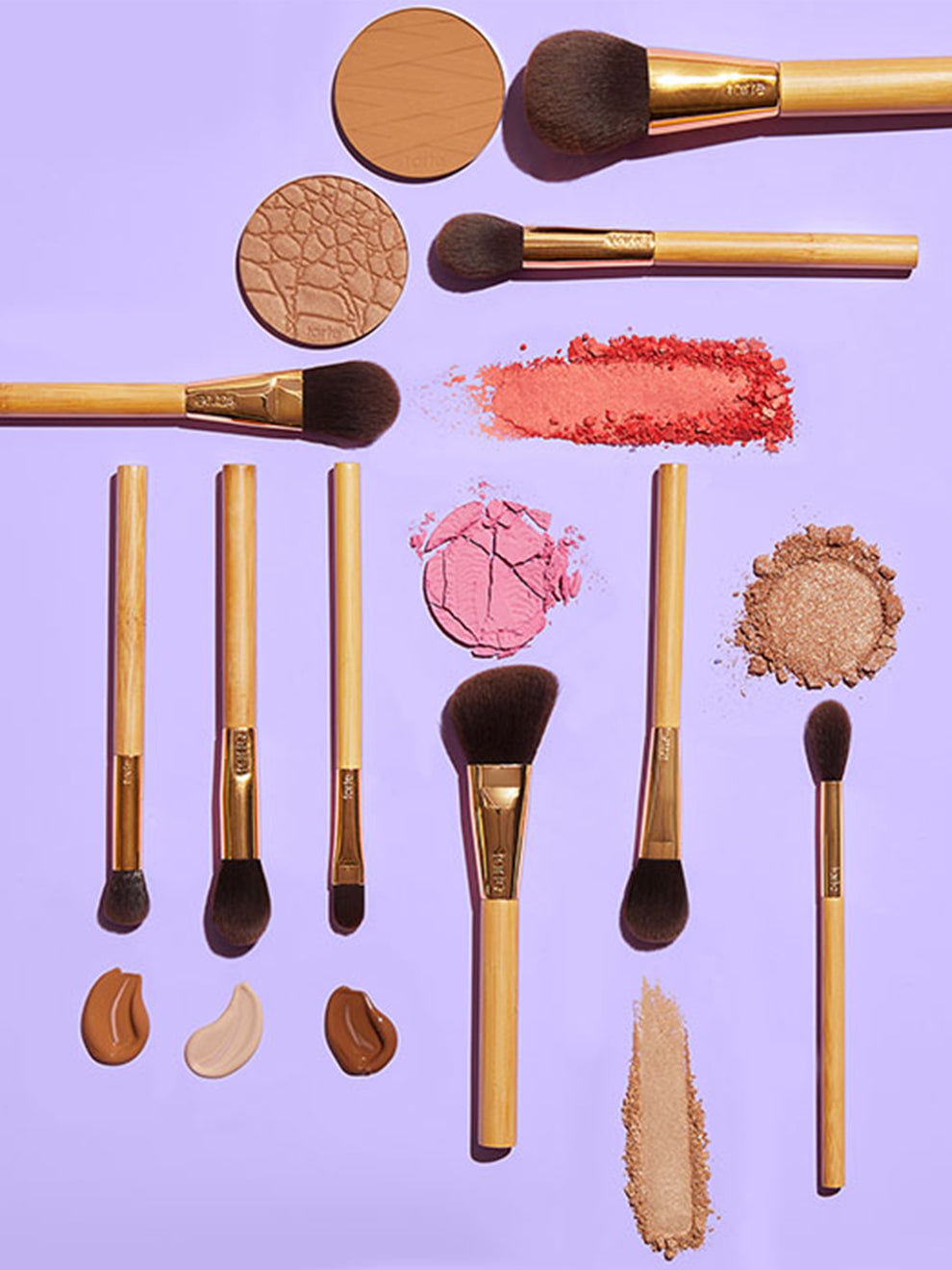 Makeup brushes lie flat on a light purple surface alongside smudged powders in shades of pink and brown. The brushes have wooden handles, and embossed tarte logos are visible.