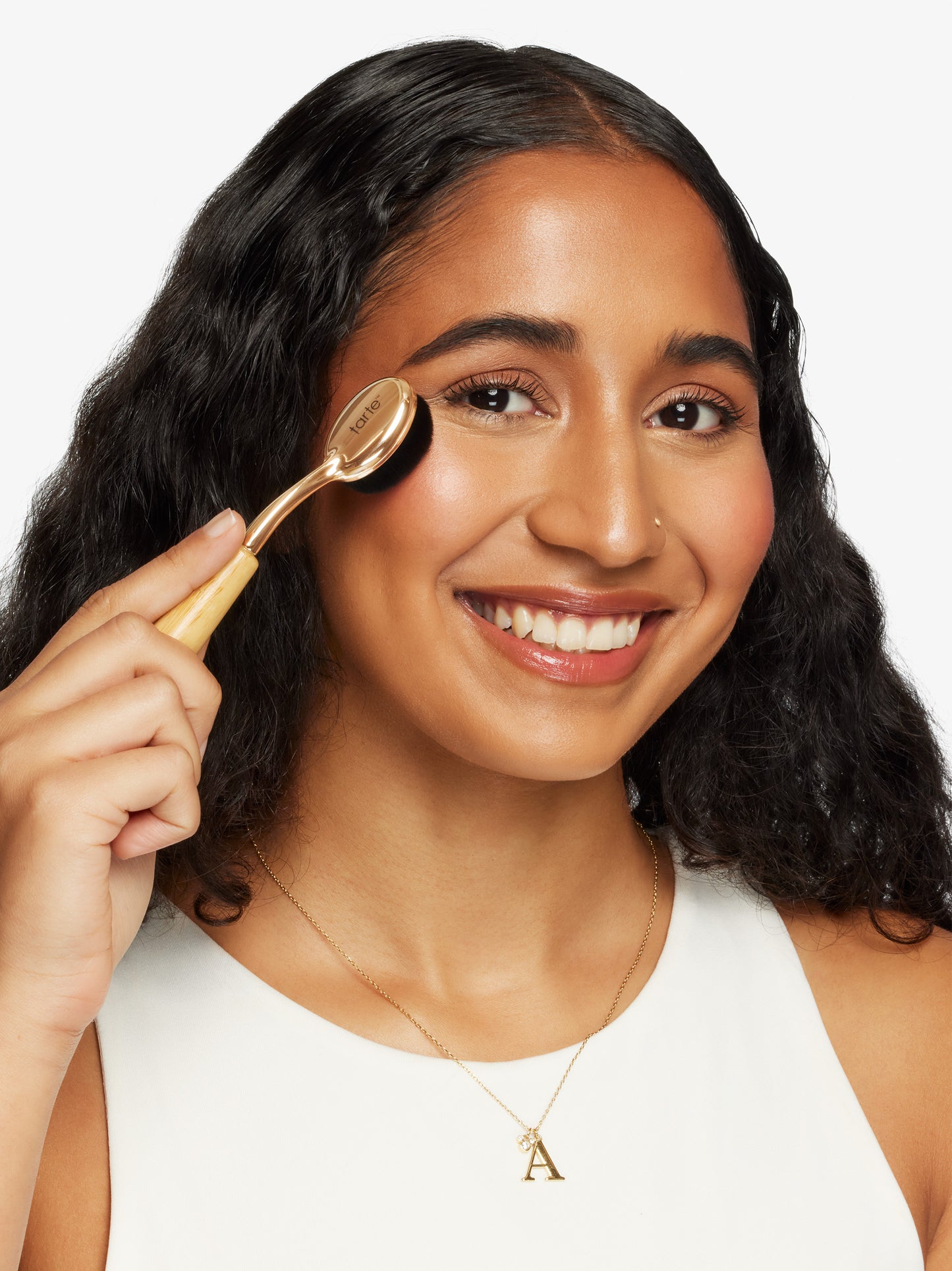 A woman uses a Tarte Cosmetics brush on her cheek, smiling. She has long, dark hair, wears a white top, and a necklace with an A pendant.