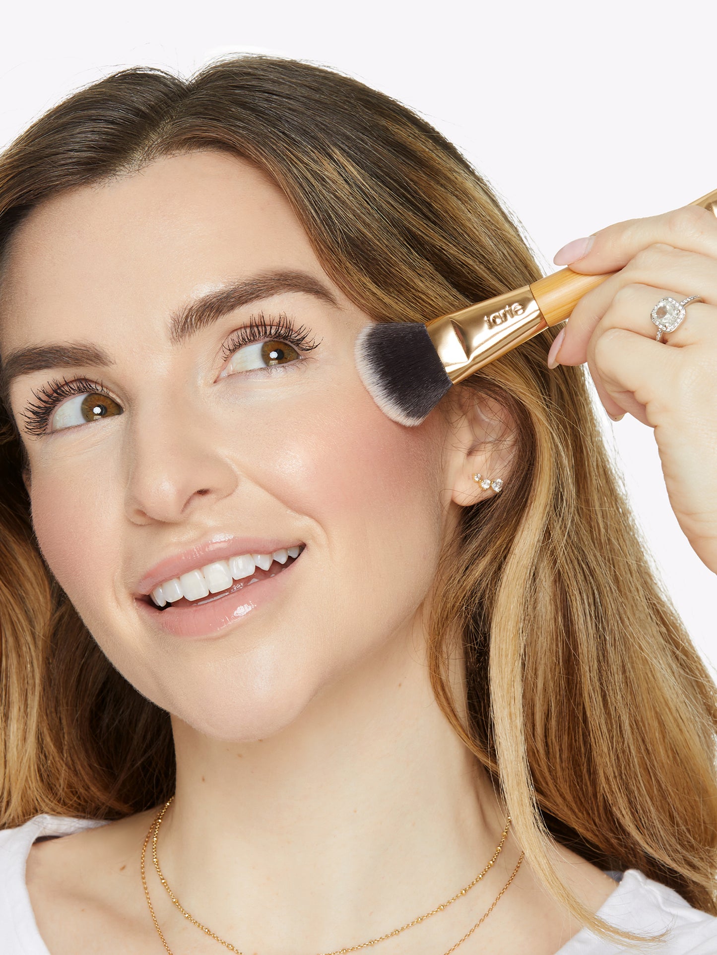 A woman applies makeup with a Tarte Cosmetics brush, smiling brightly in a well-lit setting, showcasing her earrings and necklace.