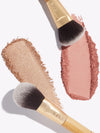 Makeup brushes spread shimmering beige and rosy pink powders on a clean white background. Each brush has a gold and wooden handle embossed with tarte.