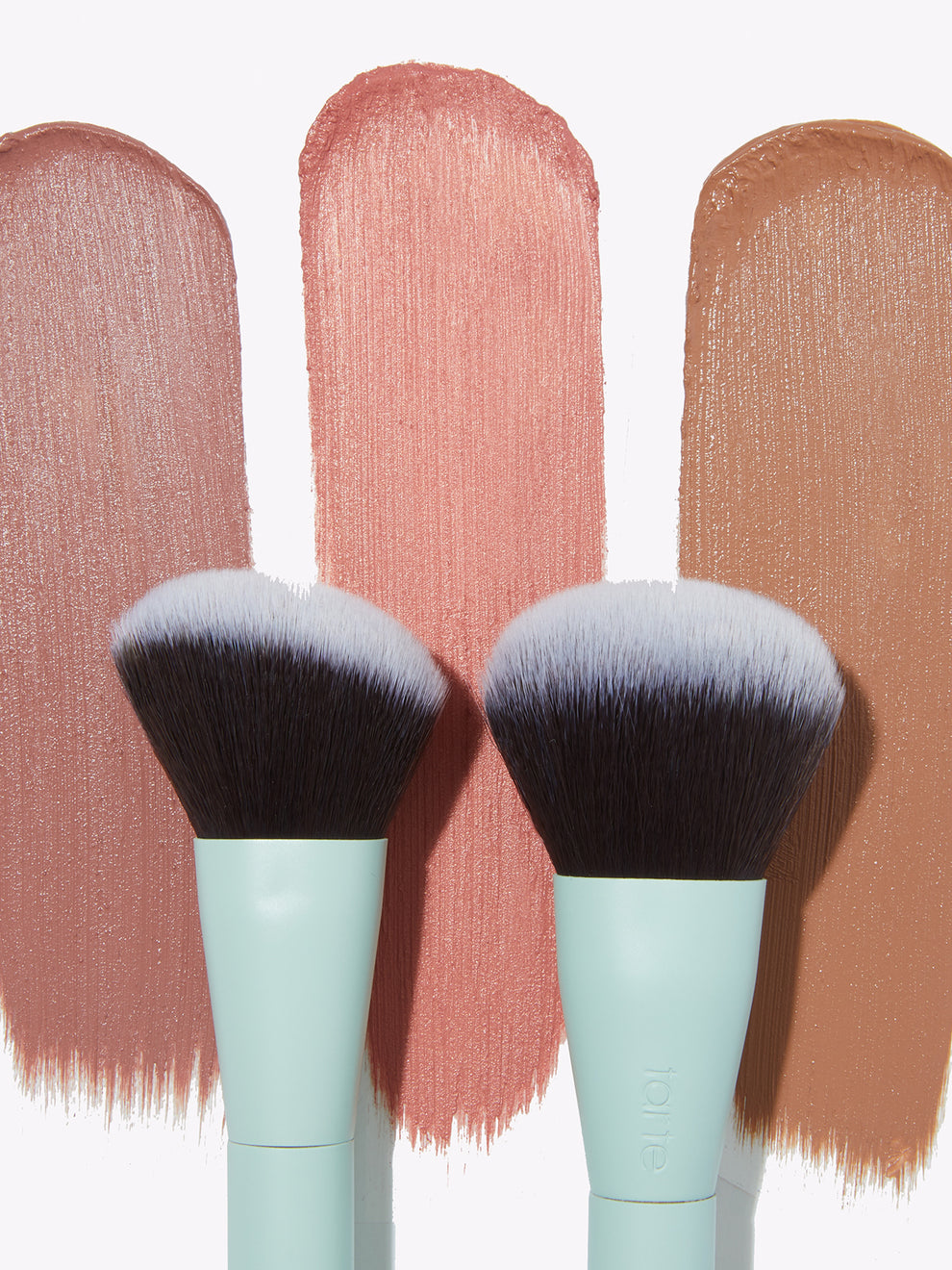 Two makeup brushes stand against swatches of creamy pinks and brown shades on a light background. Text on one brush reads tarte, indicating Tarte Cosmetics branding.