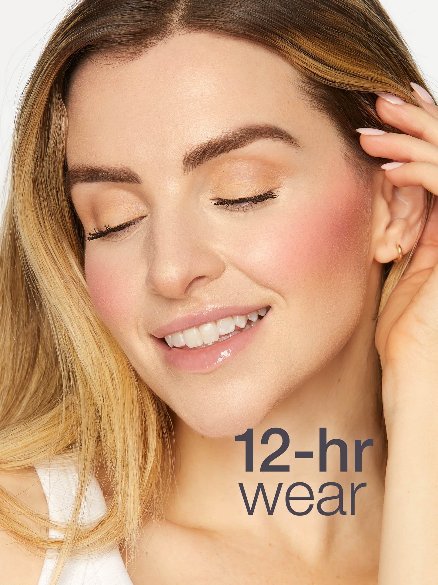 A woman with closed eyes smiles gently while touching her hair in a bright setting. Her makeup looks fresh, showcasing rosy cheeks and glossy lips. Text reads, 12-hr wear.