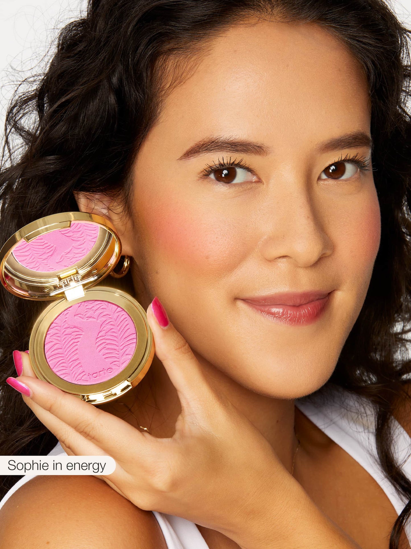 A person holds Tarte Cosmetics blush in a gold compact, showcasing vibrant pink color embossed with an intricate design. Text: Sophie in energy. The backdrop is a simple white setting.