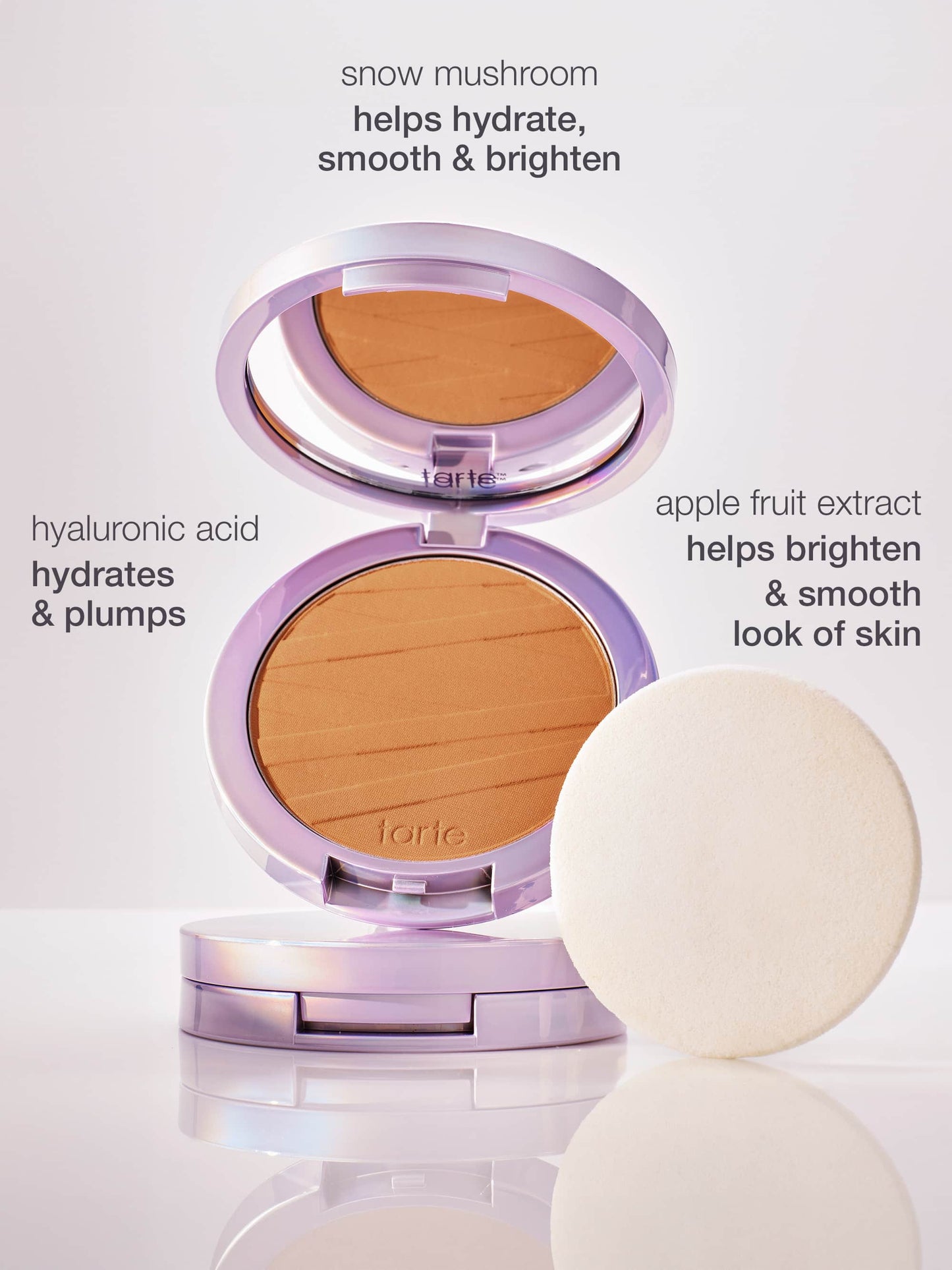 A Tarte Cosmetics powder compact is open, displaying its product alongside a white application sponge. Text highlights ingredients: snow mushroom helps hydrate, smooth & brighten, hyaluronic acid hydrates & plumps, apple fruit extract helps brighten & smooth look of skin. The background is a soft, neutral surface.