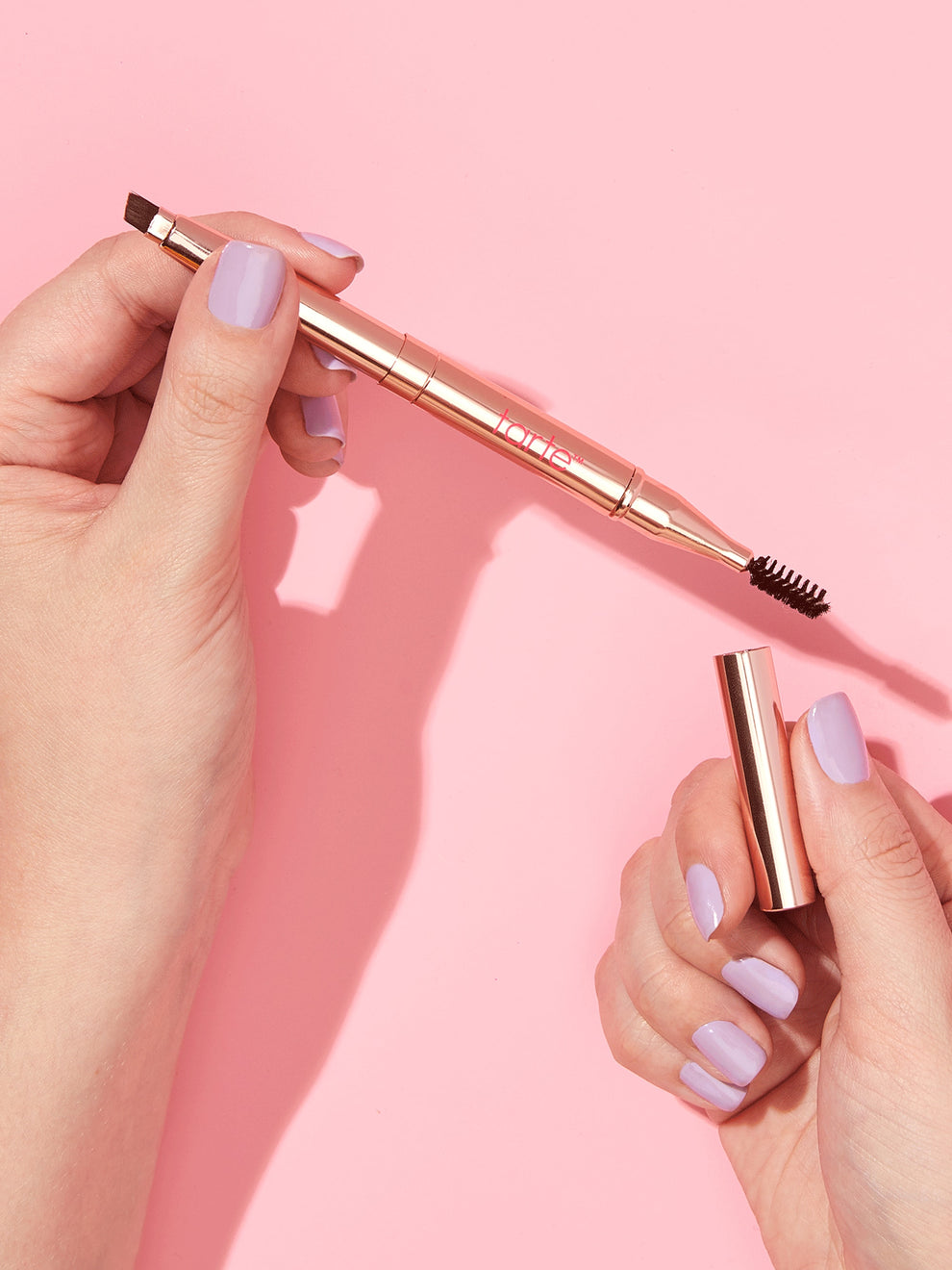 A dual-ended Tarte Cosmetics eyebrow tool with a brush and spoolie is held by two hands over a pink background. The hands have light purple nail polish.