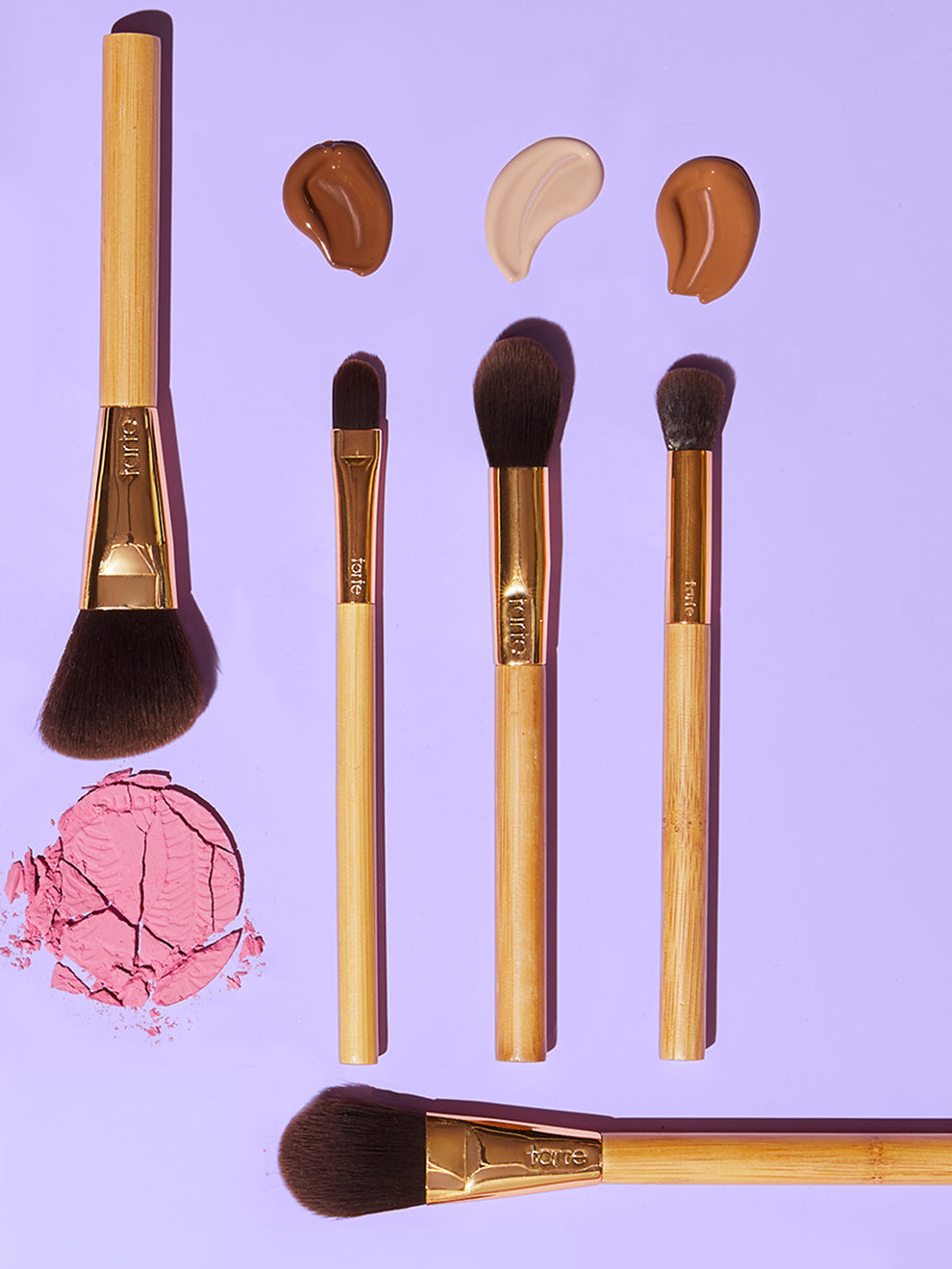 Five Tarte Cosmetics brushes are arranged on a purple surface, accompanied by smears of three foundation shades and a crushed pink blush.