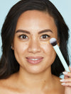 Woman applying makeup with a Tarte Cosmetics brush, gently blending under-eye concealer in a close-up shot against a plain blue background.