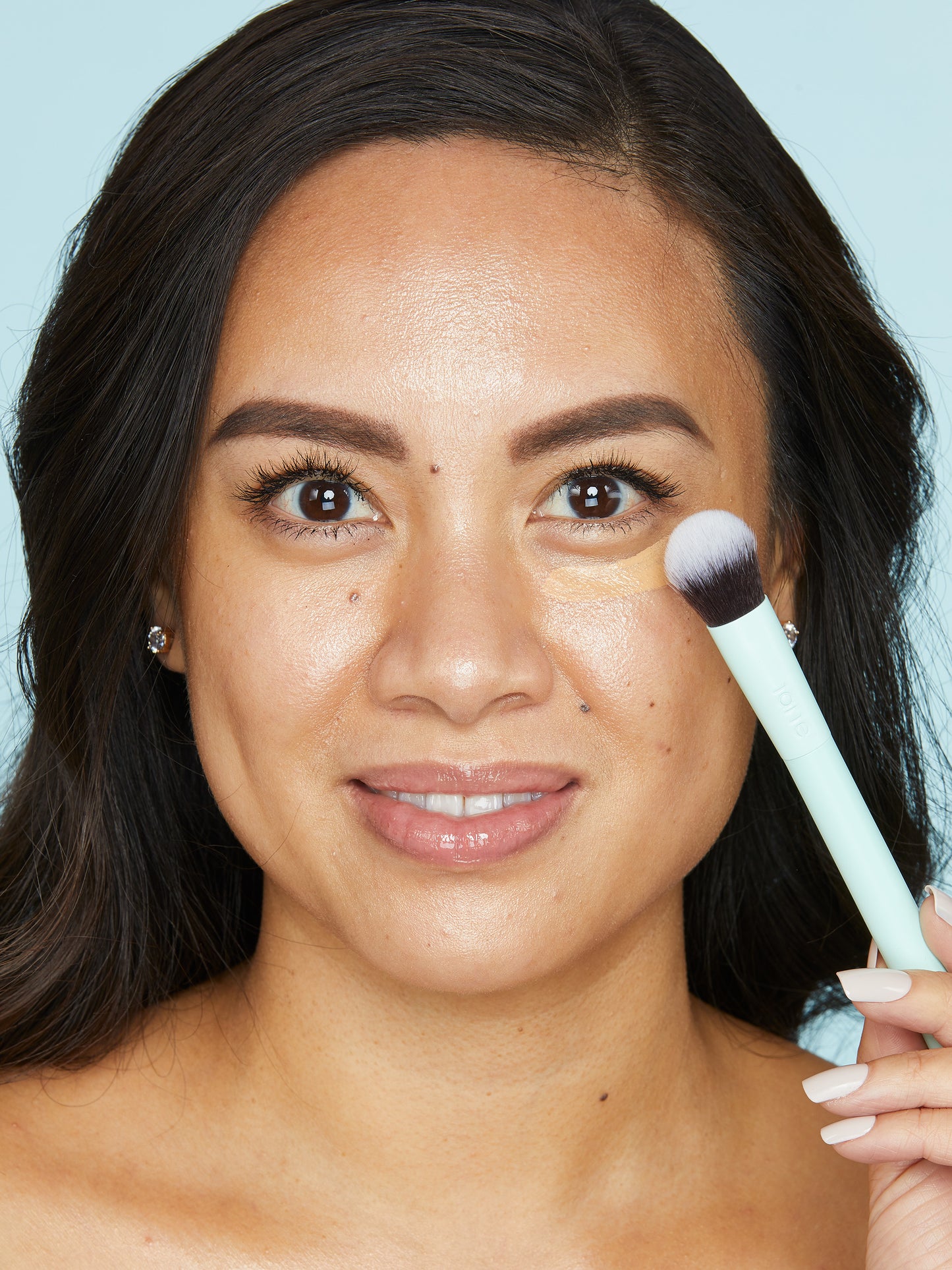 Woman applying makeup with a Tarte Cosmetics brush, gently blending under-eye concealer in a close-up shot against a plain blue background.