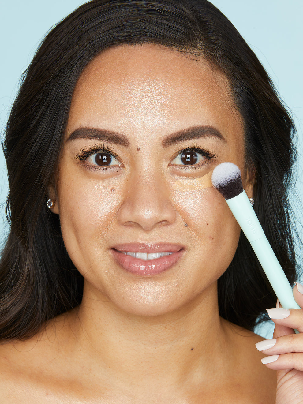 Woman applying makeup with a Tarte Cosmetics brush, gently blending under-eye concealer in a close-up shot against a plain blue background.