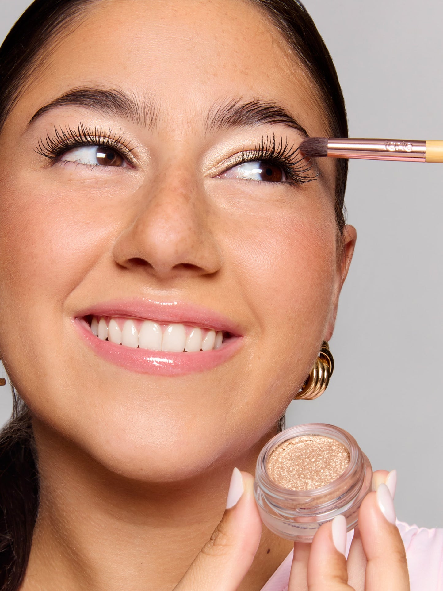 A person smiles while applying shimmery eyeshadow with a brush. They hold a small jar of glittery makeup. The background is a plain, light gray.
