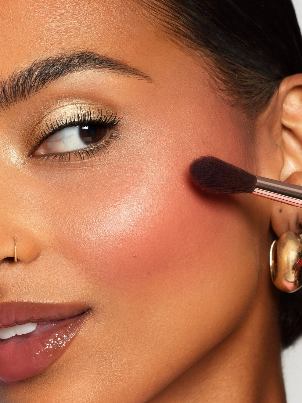A person is applying blush with a makeup brush to their cheek. The contents of the photo includes a woman wearing gold eyeshadow, glossy lips, hoop earrings, and a nose ring, highlighting a polished makeup look.