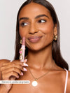 A person holds Tarte Cosmetics' lipstick close to their face, smiling with shiny lips, wearing a gold necklace and hoop earrings. Text: Remmi in vanilla icing balm.