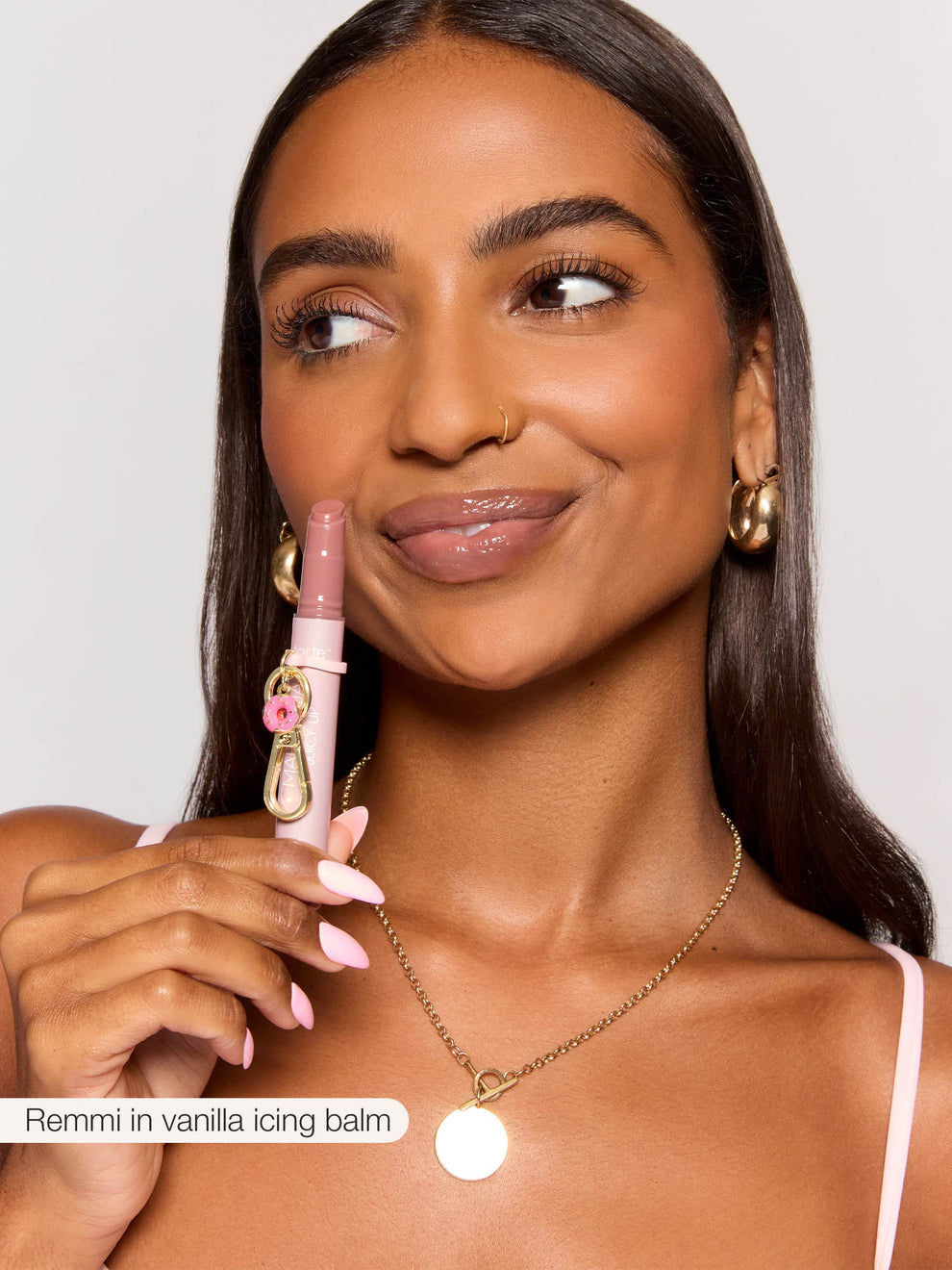 A person holds Tarte Cosmetics' lipstick close to their face, smiling with shiny lips, wearing a gold necklace and hoop earrings. Text: Remmi in vanilla icing balm.