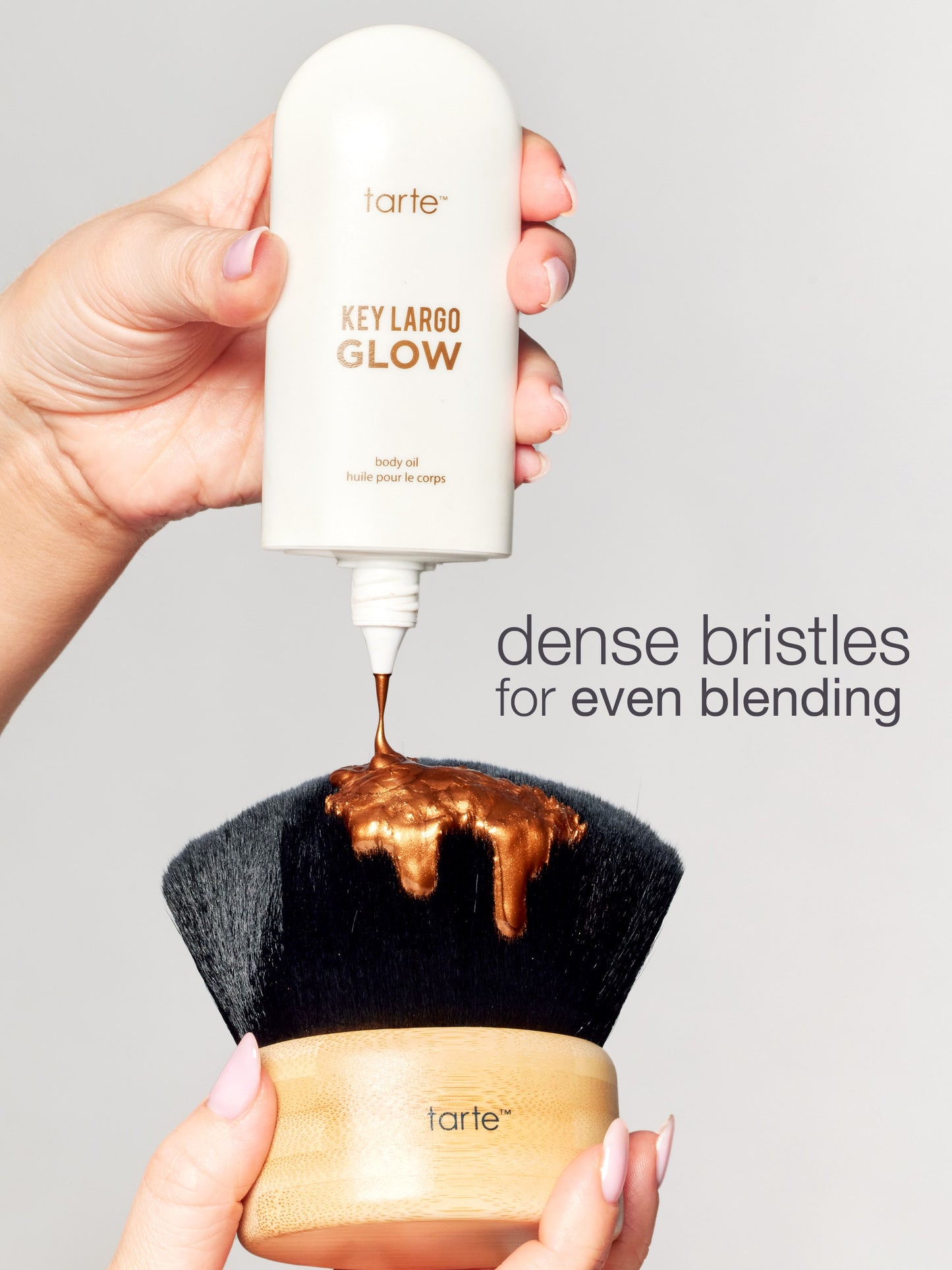 Tarte makeup brush with brown powder being applied, held by a hand against a gray background.