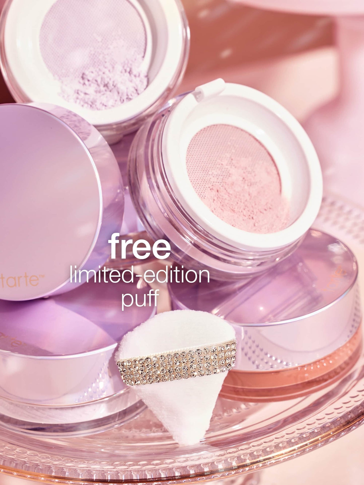 Tarte Cosmetics translucent powder containers are open, revealing pastel powders on a glass tray. A small, soft puff with a rhinestone band is displayed. Text reads, free limited-edition puff.