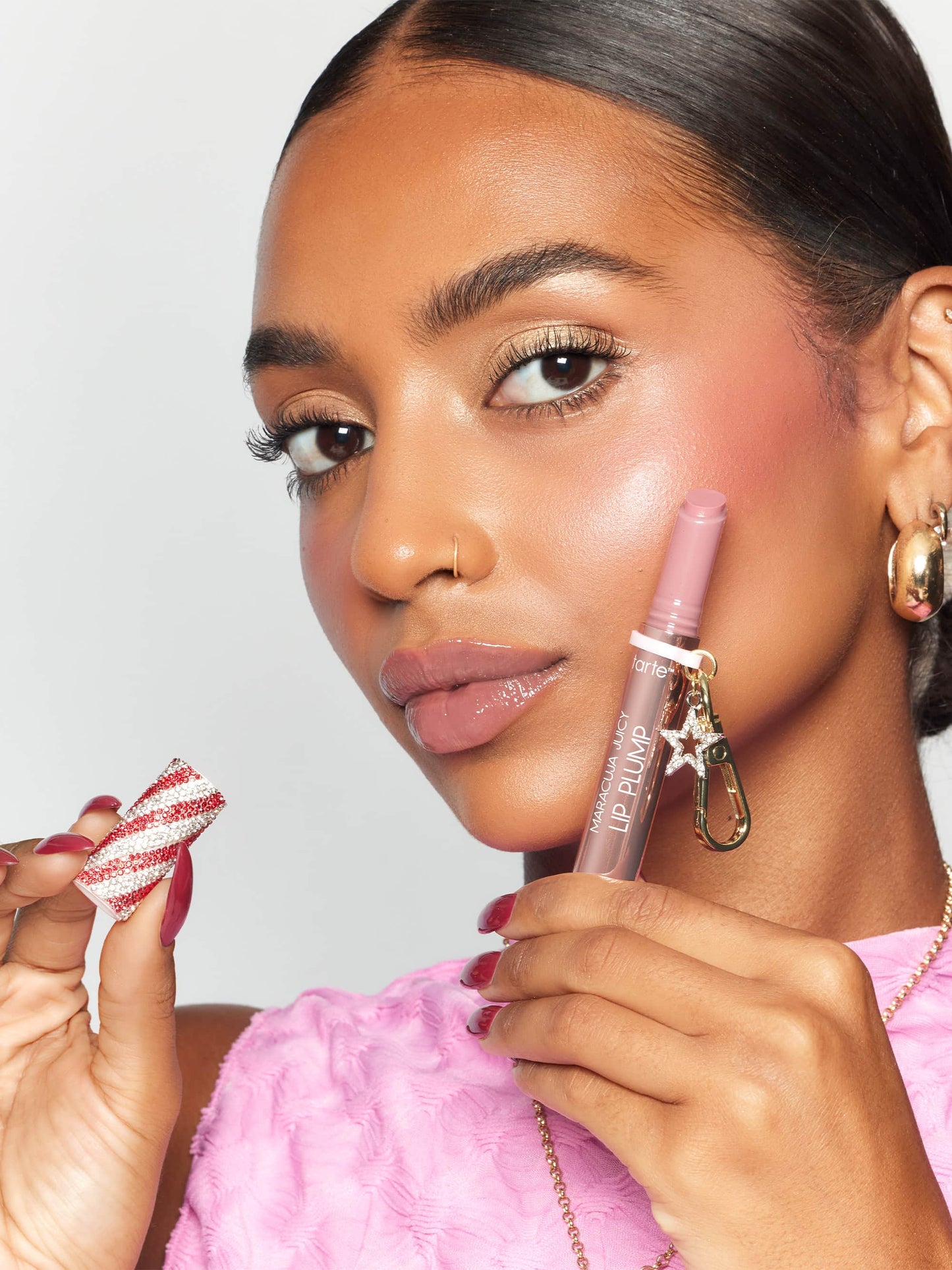 A person holds a Tarte Cosmetics Maracuja Juicy Lip Plump tube near their lips, wearing a pink textured top. Their other hand holds a decorative red-and-white striped cap.
