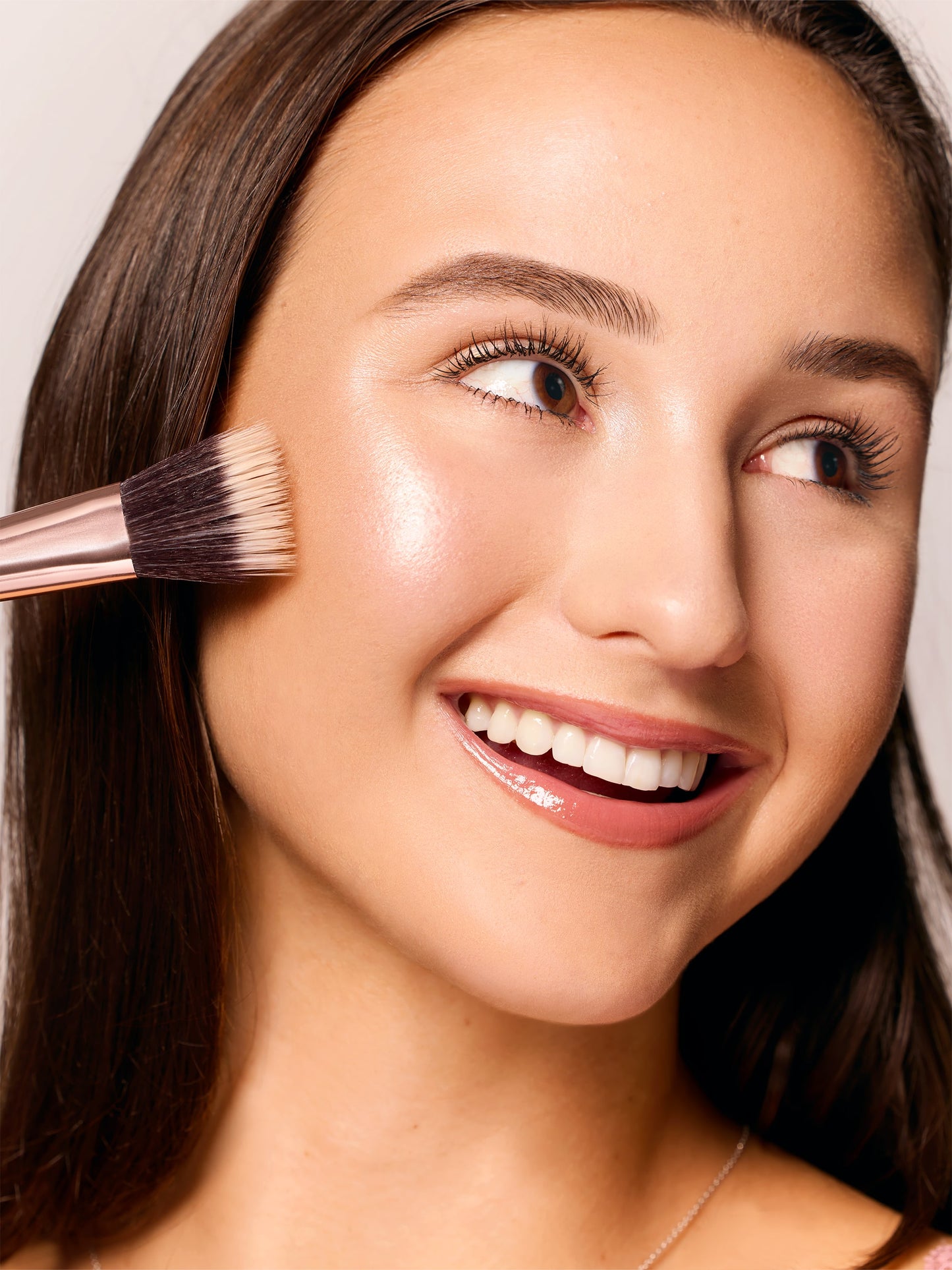 A makeup brush applies highlighter to a smiling person's cheek, accentuating their glowing complexion in a close-up shot. The background is softly blurred, highlighting the focus on the face.