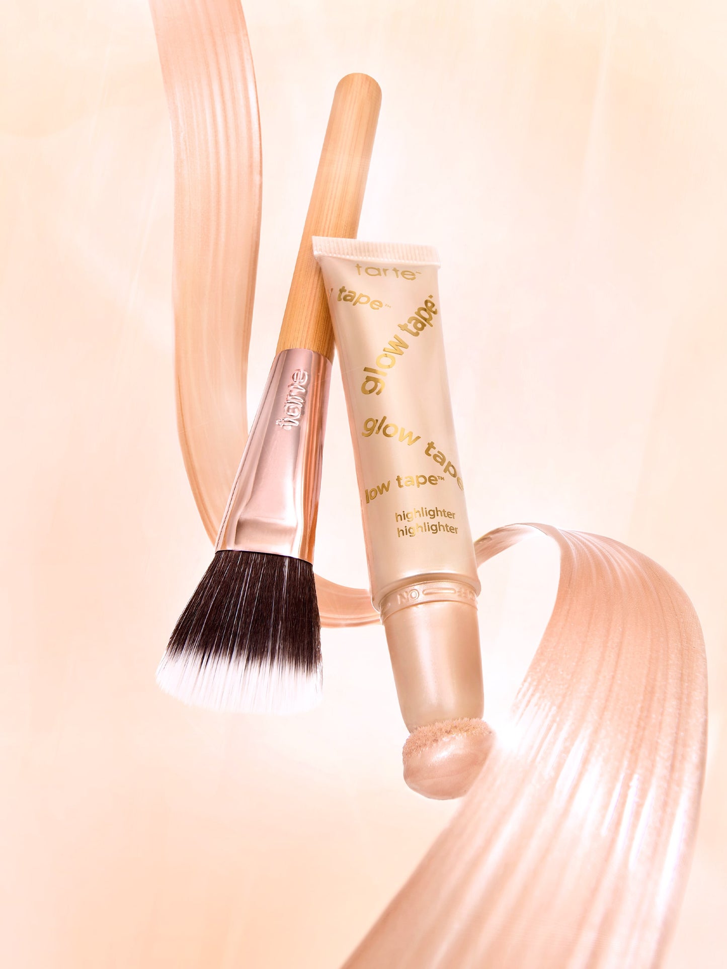 A Tarte Cosmetics brush and glow tape highlighter are placed on a light peach background, accompanied by swirling strokes of highlighter product. Text on tube: tarte glow tape glow tape highlighter highlighter.
