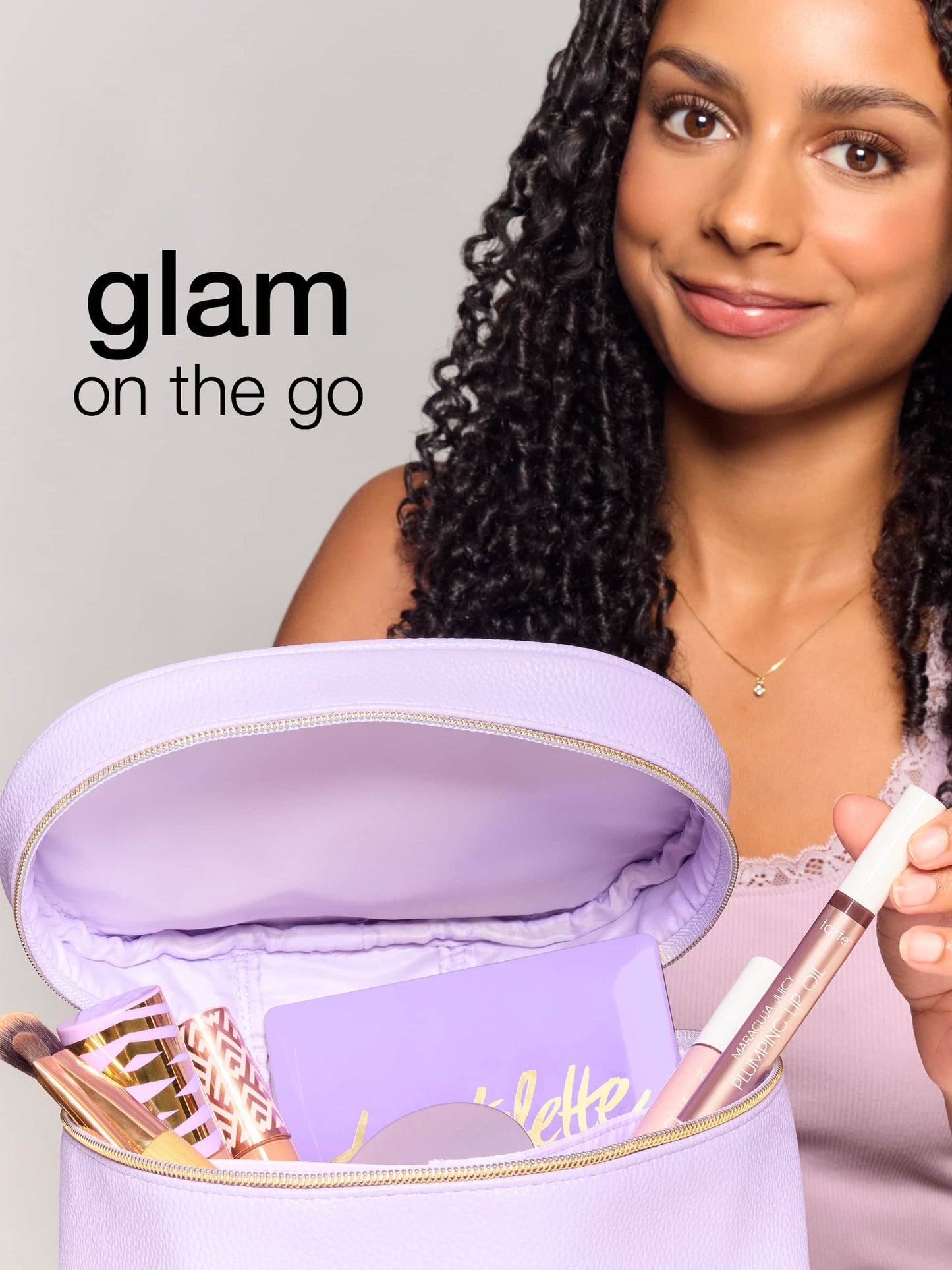 A woman smiles while holding an open lavender makeup bag containing Tarte Cosmetics products, including a tartelette palette and plumping lip oil. Text reads, glam on the go.