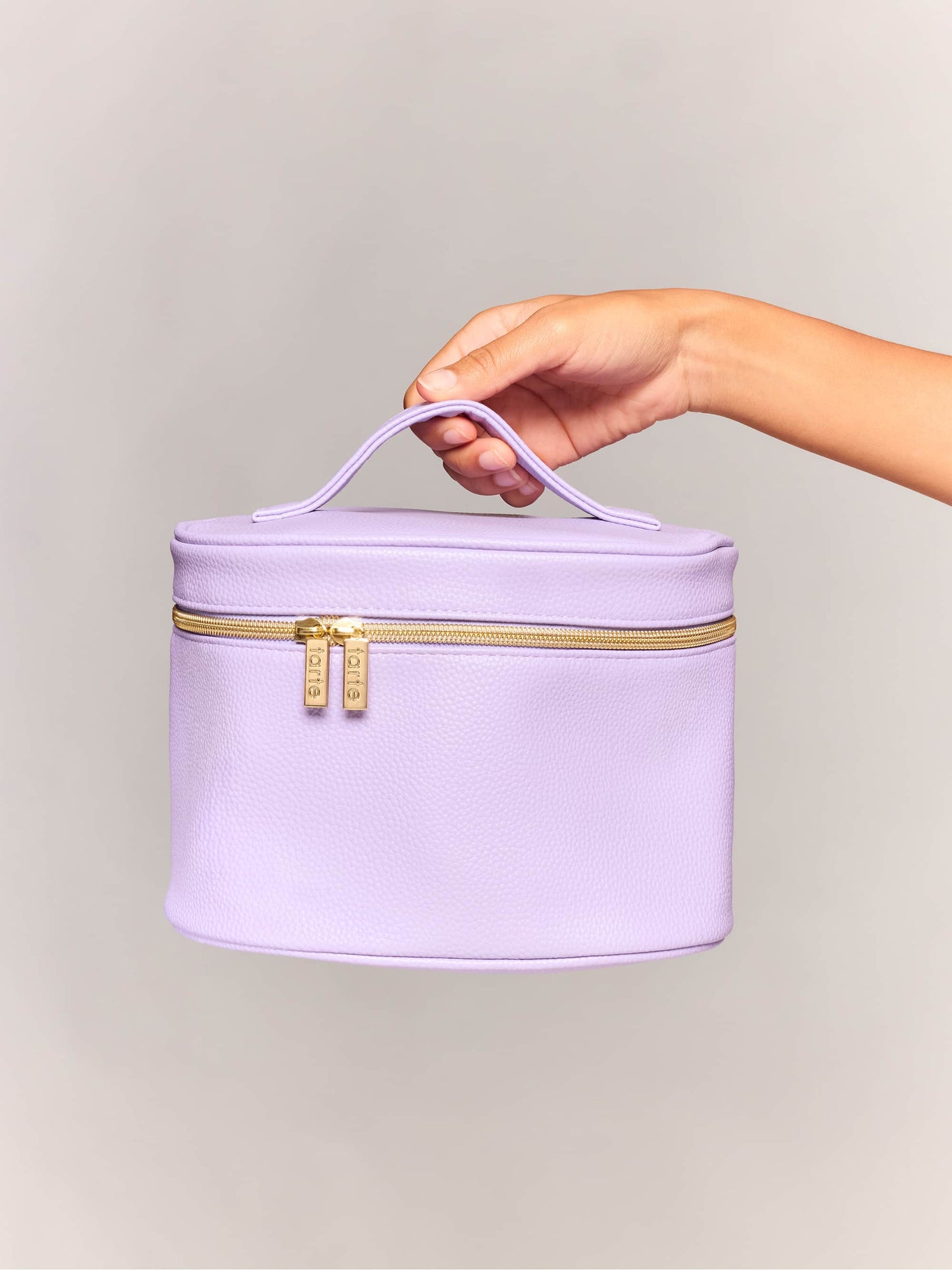 A lavender makeup bag with a handle is held by a hand, showcasing a zippered closure with two gold zippers labeled Tarte, against a plain, light gray background.