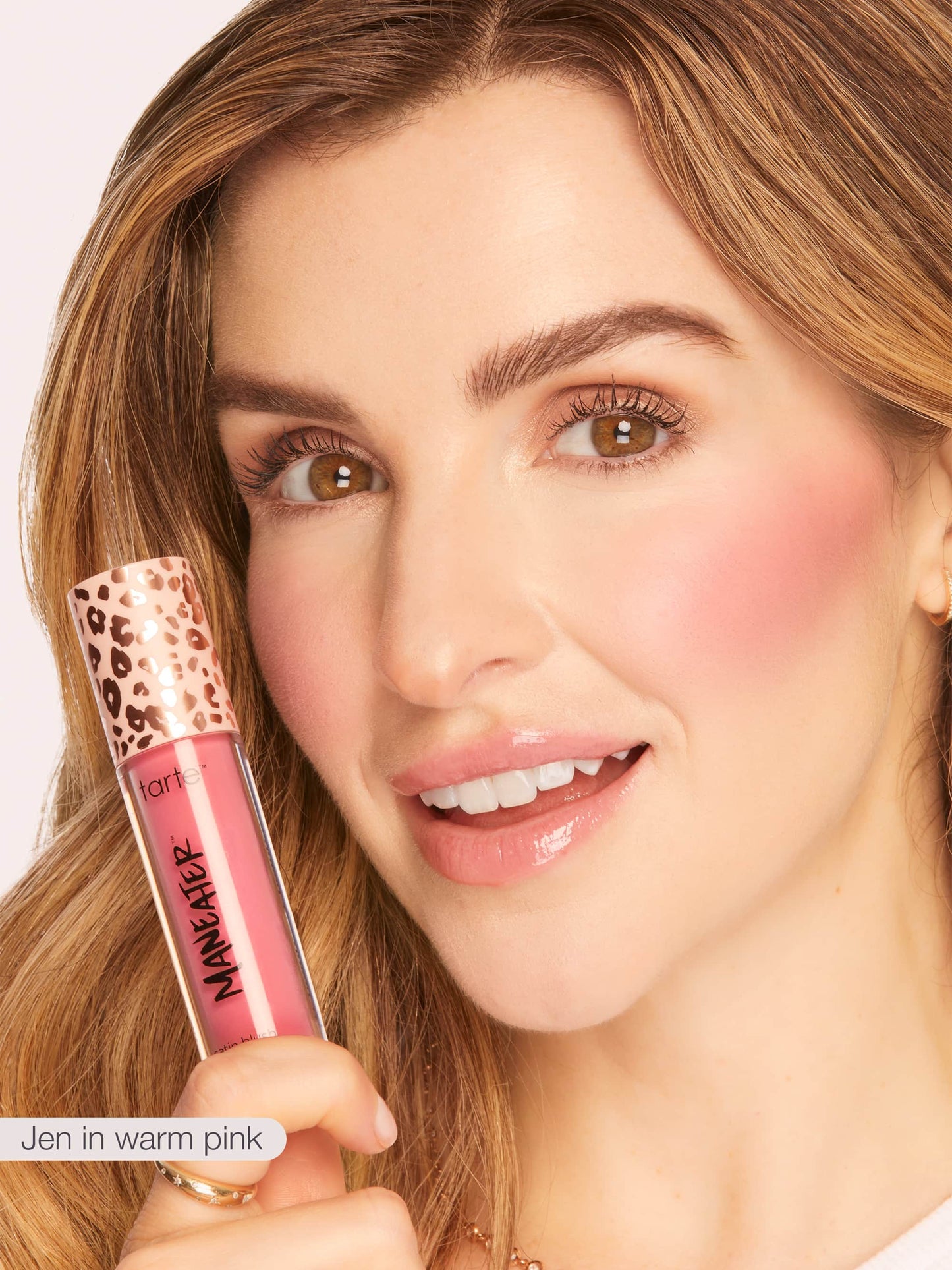 A woman holds Tarte Cosmetics Maneater lip gloss in warm pink near her face, smiling against a light background. Text reads: Jen in warm pink.