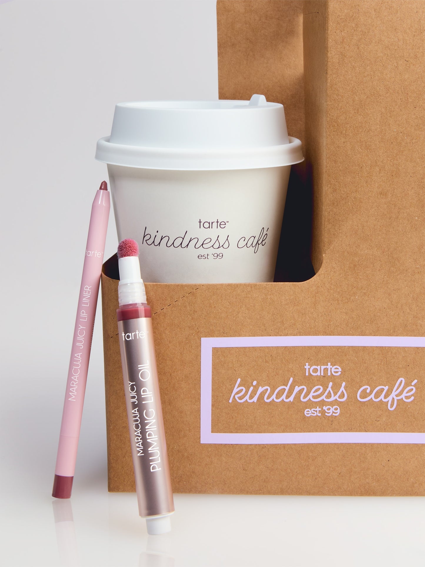 A Tarte Cosmetics lip liner and plumping lip oil are displayed beside a coffee cup and brown paper bag labeled tarte kindness café est '99 on a reflective surface.