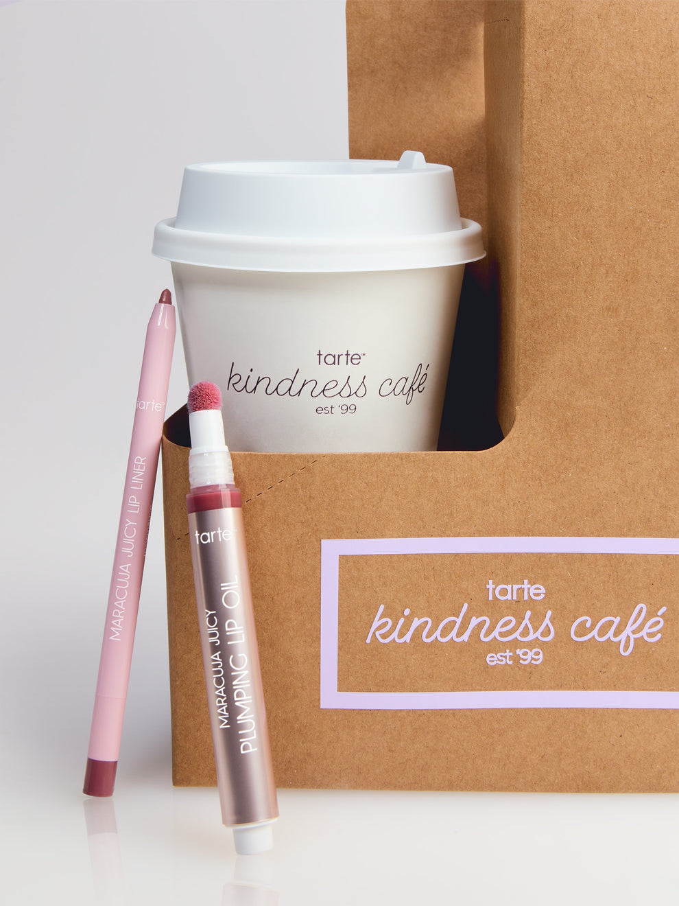 A Tarte Cosmetics lip liner and plumping lip oil are displayed beside a coffee cup and brown paper bag labeled tarte kindness café est '99 on a reflective surface.