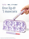 A hand holds a limited edition tarte cosmetics x roller rabbit collaboration tartelette tubing mascara above a white zip pouch printed with purple cartoon monkeys. A matching lip item lies in front of the pouch.