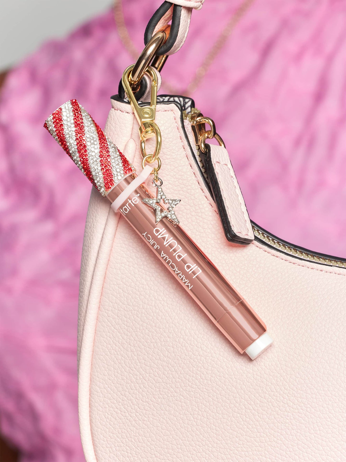 Lip gloss tube with a red and white glitter cap attached to a pink handbag by a gold clasp, featuring a small sparkling star charm. Background shows a pink fabric. Text: tarte MARACUJA JUICY LIP PLUMP.