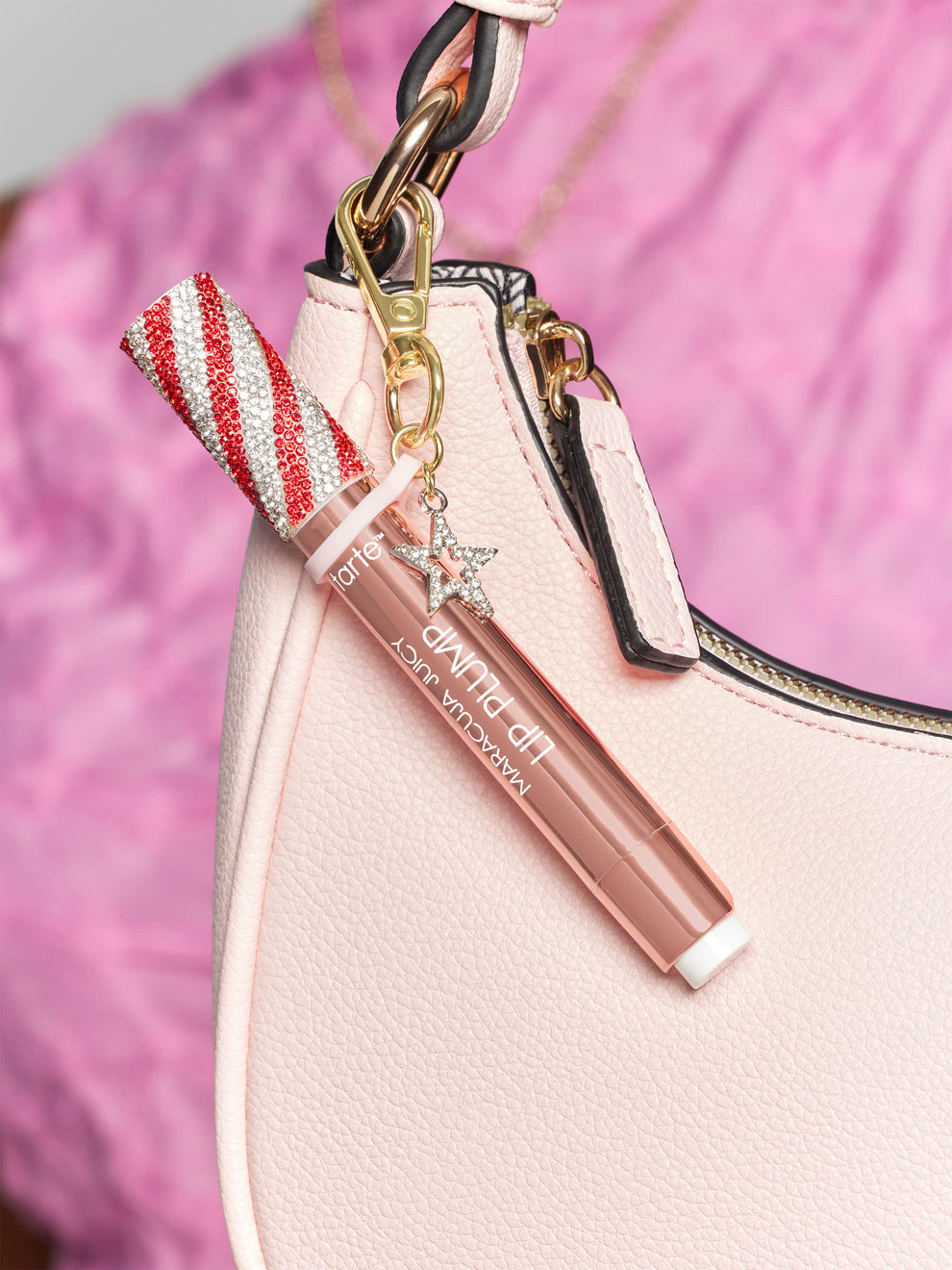 Lip gloss tube with a red and white glitter cap attached to a pink handbag by a gold clasp, featuring a small sparkling star charm. Background shows a pink fabric. Text: tarte MARACUJA JUICY LIP PLUMP.