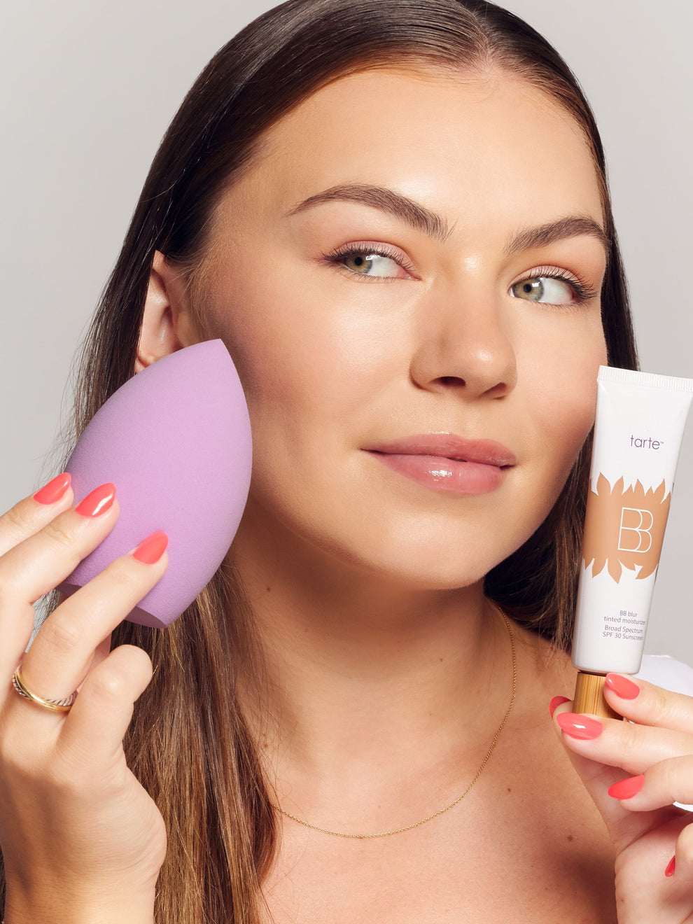 Woman holding a purple makeup sponge and a Tarte BB cream.